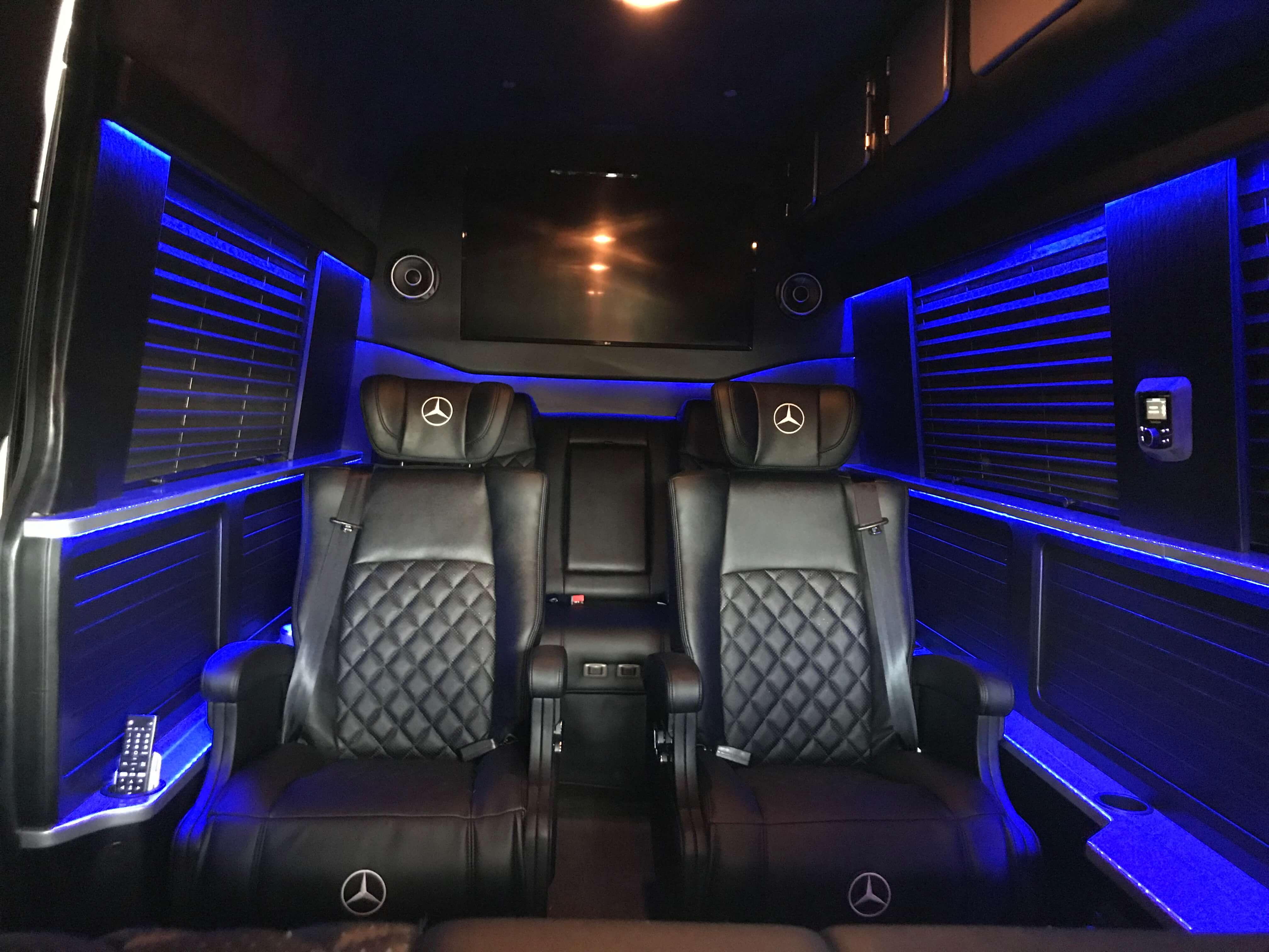 Mercedes-branded captain chairs in a blue LED-lit van conversion with wall-mounted entertainment screen and integrated speakers